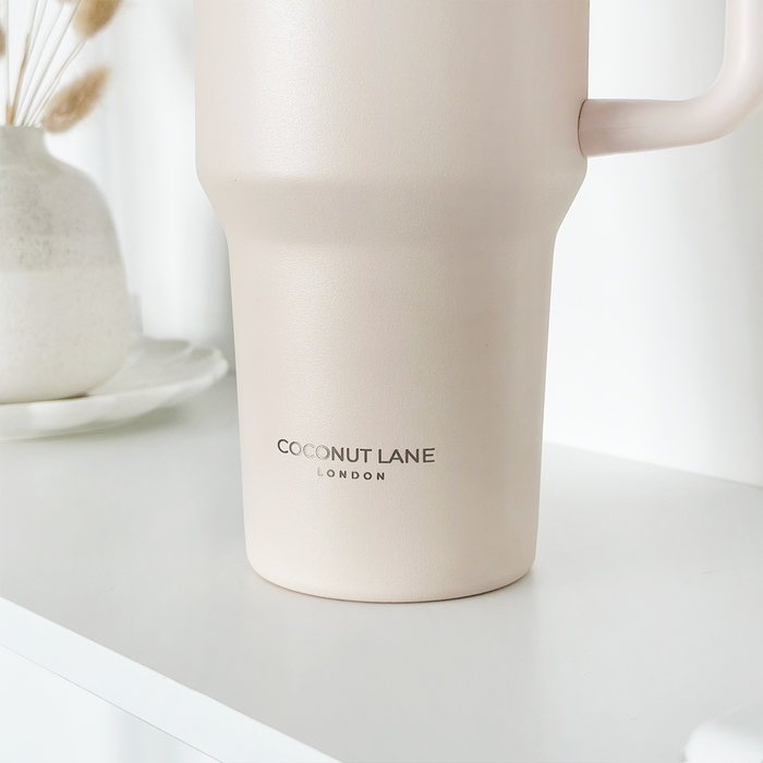Marshmallow pink Cocopup London stainless steel tumbler on white surface beside a vase of dried flowers.