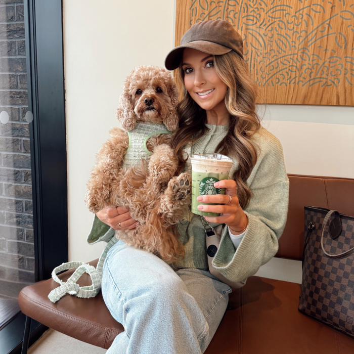 Woman holds a fluffy brown dog wearing a green textured tweed Cocopup London harness, holding a green Starbucks drink.