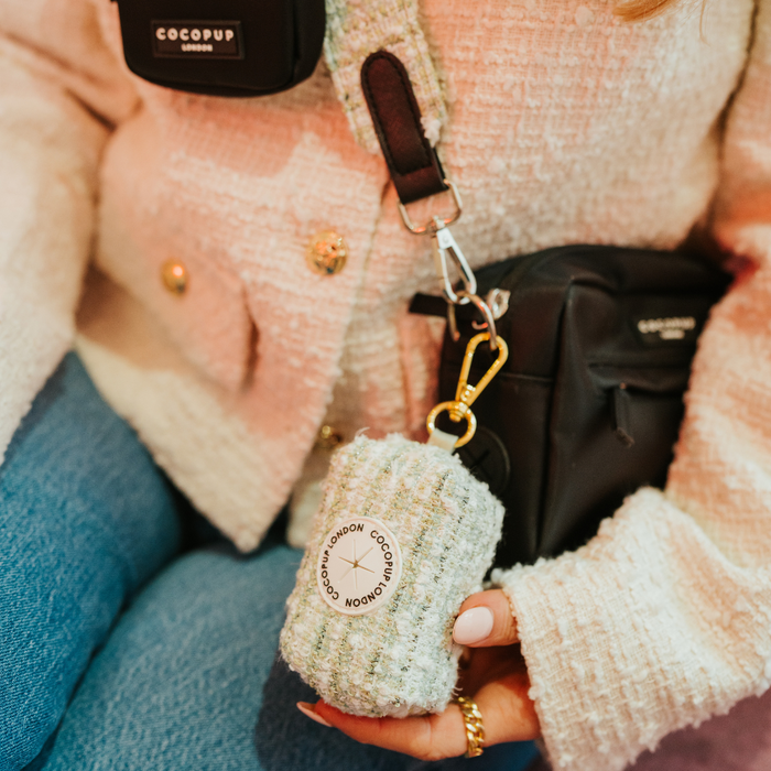 Matcha tweed Cocopup London poop bag holder clipped to a black crossbody bag, held by someone in a cream jacket.