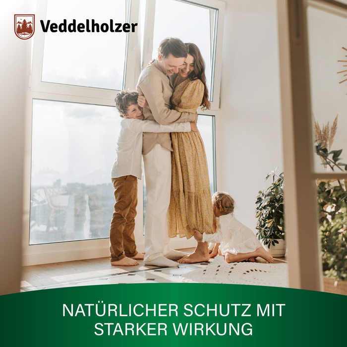 Eltern umarmen sich am hellen Fenster, der Sohn schmiegt sich an, die kleine Tochter spielt auf dem Teppich