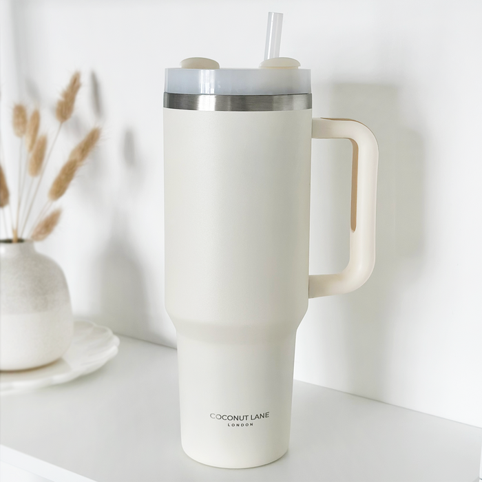 Stainless steel tumbler with handle and straw in a milky coconut finish, sitting on a white shelf.