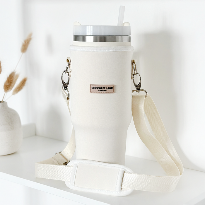 Creamy white tumbler carry case with cross-body strap and straw lid, displayed on a white shelf.