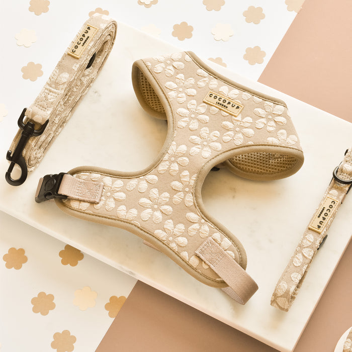 A matching dog harness, leash, and collar set in a luxe mocha color with a white floral print. The adjustable neck harness features the brand name "Cocopup London" on a small tag. These bundled pet accessories are displayed on a white marble surface with floral decorations in the background.