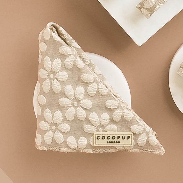 A light brown Luxe Bandana - Mocha Flower from Cocopup London, featuring white floral patterns and a tag that reads "COCOPUP LONDON," is displayed on a neutral background. This elegant piece of pup fashion is neatly folded into a triangle shape.