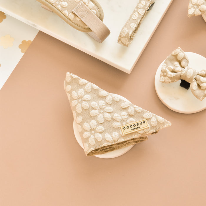A collection of beige dog accessories with white floral patterns is displayed on a marble slab and beige background. Items include a folded Cocopup London Luxe Bandana - Mocha Flower, a bow tie, and parts of a leash and other pup fashion accessories.