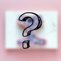 A large, bold question mark is centered over a blurred background with soft pastel colors evoking light pink and white hues. The overall image from Cocopup London captures a sense of curiosity and mystery, reminiscent of the hidden secrets in the Mystery Adjustable Neck Harness.