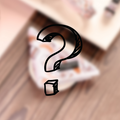 A large, black question mark with a small cube at its base is centered on a blurred background of soft, earthy tones. The background seems to suggest Cocopup London's stylish Mystery Bandana on a wooden surface, but it is not clearly visible.