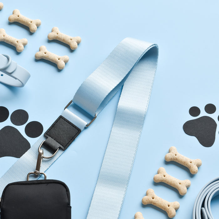 A light blue dog leash, a roll of waste bags, a black waste bag holder, and a light blue collar are arranged on a blue surface. Several bone-shaped dog treats and black paw print cutouts are scattered around the accessories, making it perfect for pairing with your Cocopup London's NAKD Bag Strap - Bubblegum.