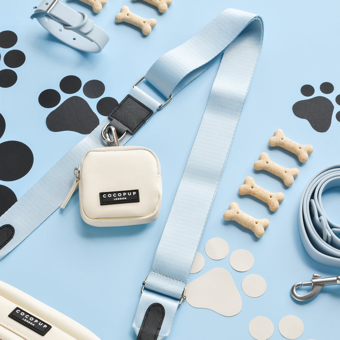 A blue background showcases various dog accessories, including a light blue leash, collar, and waste bag dispenser. Among bone-shaped dog treats lies a small white pouch labeled "Cocopup London," featuring the versatile Cocopup Dog Walking Bag with an optional NAKD Bag Strap - Bubblegum. Black and white paw prints are scattered around.