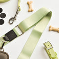 Assorted pet accessories arranged on a white surface, featuring the Cocopup London NAKD Bag Strap in Matcha with a metal hook, dog bone-shaped treats, a collar with a buckle, and black circular stickers. The collection also includes stylish items compatible with the Cocopup Dog Walking Bag.