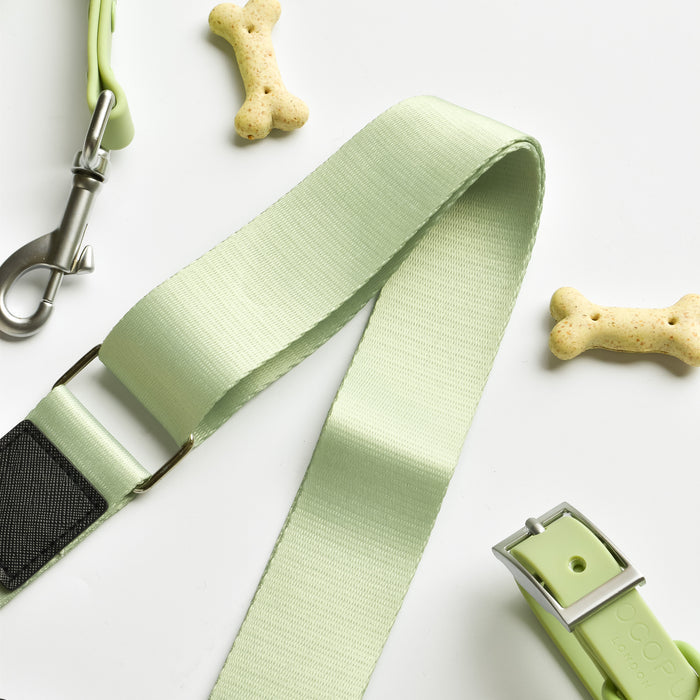 A light green dog collar and leash set, featuring sage-coloured straps with black accents, is displayed against a white background. Scattered around the leash and collar are bone-shaped dog biscuits. This set pairs perfectly with the Cocopup London NAKD Bag Strap - Matcha from the NAKD Matcha collection.