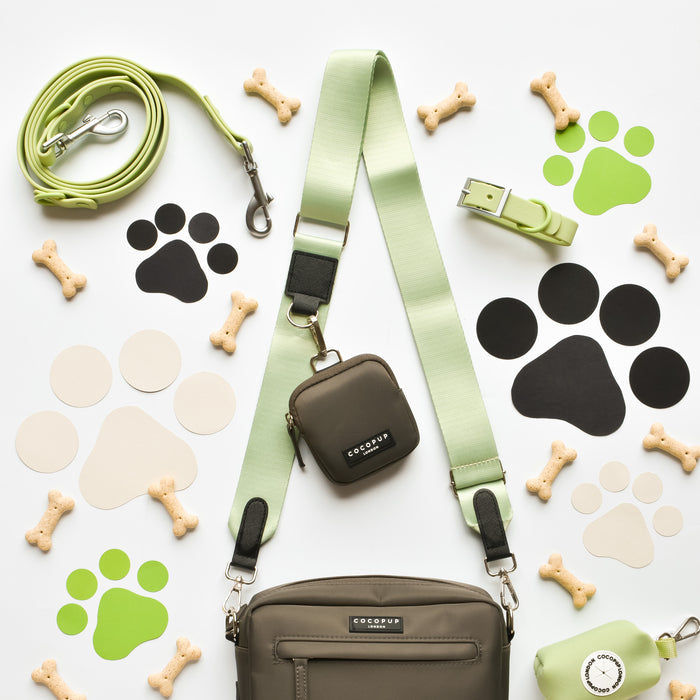 A flat lay of pet accessories, including a green leash, scattered dog bone treats, paw print stickers in green, beige, and black, a small green bag from the NAKD Matcha Collection featuring the NAKD Bag Strap - Matcha by Cocopup London, a matching collar with a sage-colored strap, and a green waste bag holder. Also included is the Cocopup Dog Walking Bag with an attached pouch by Cocopup London.