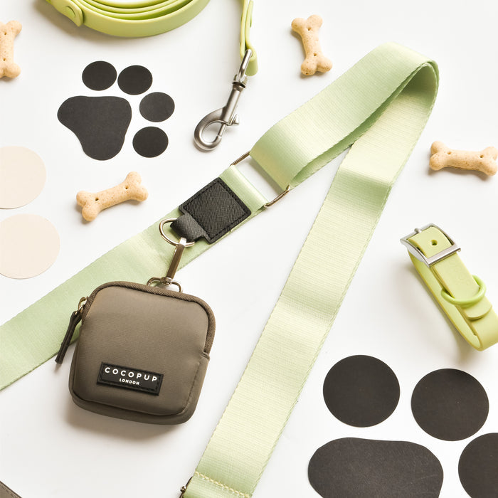 A set of dog accessories arranged on a white surface, including a NAKD Bag Strap - Matcha with a sage-colored strap, a matching buckle collar, a small green pouch labeled "Cocopup London," several bone-shaped dog biscuits, and black paw print cutouts.