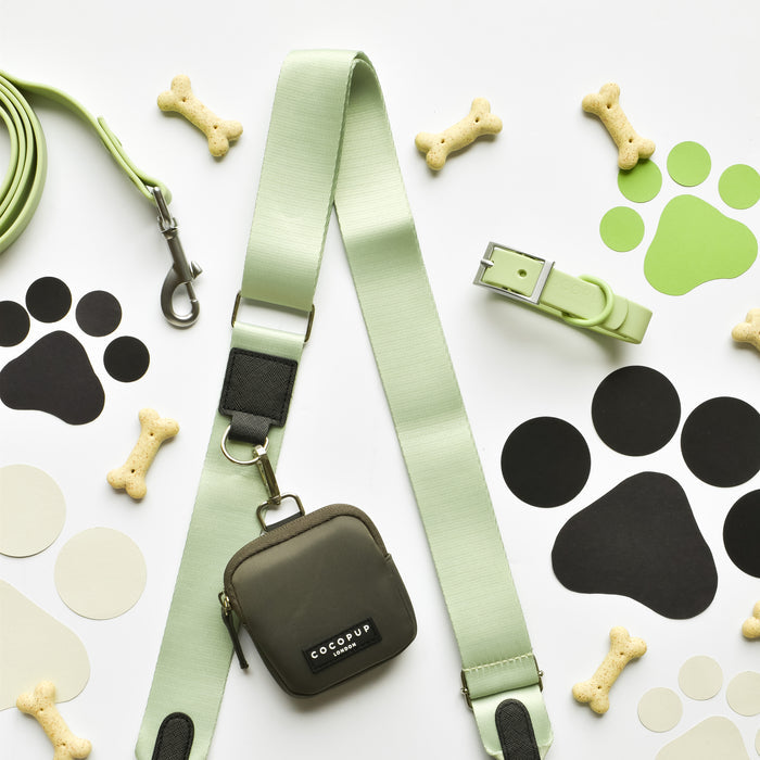 A neatly arranged set of dog accessories includes a green leash, collar, and poop bag holder from the NAKD Matcha collection. Surrounding them are bone-shaped dog treats, black and green paw print cutouts, decorative circles on a white background, and a Cocopup London Dog Walking Bag featuring the NAKD Bag Strap in Matcha.