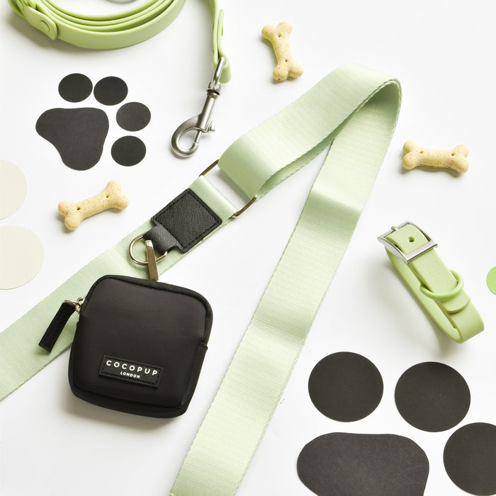 A leash and collar set in light green, featuring a sage-colored strap, is complemented by a small black zipper pouch labeled "Cocopup London." These items are part of the NAKD Bag Strap - Matcha collection and are displayed with bone-shaped dog treats and black-and-white paw print cutouts.