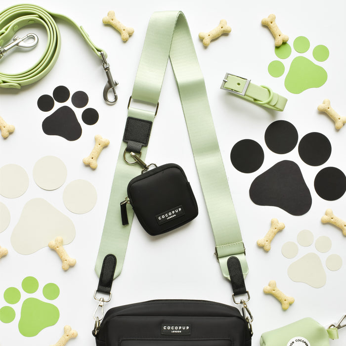 A flat lay image showcases the Cocopup London NAKD Matcha collection, featuring dog accessories including a mint green leash, harness, and collar. Alongside these items are a black poop bag holder and matching pouch. Dog biscuits and paw print cutouts in black, green, and beige are scattered across a white background.