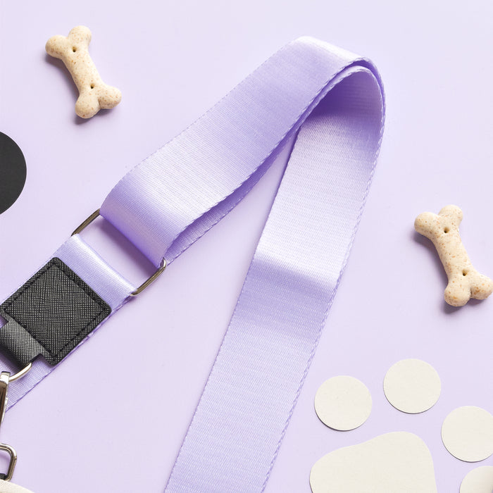 A light purple bag strap from the Cocopup London NAKD Bag Strap - Parma Violets collection is arranged on a lilac background. Surrounding the strap are several dog-themed items, including two bone-shaped biscuits and a few white circular cutouts resembling paw prints.