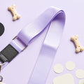 A light purple bag strap from the Cocopup London NAKD Bag Strap - Parma Violets collection is arranged on a lilac background. Surrounding the strap are several dog-themed items, including two bone-shaped biscuits and a few white circular cutouts resembling paw prints.