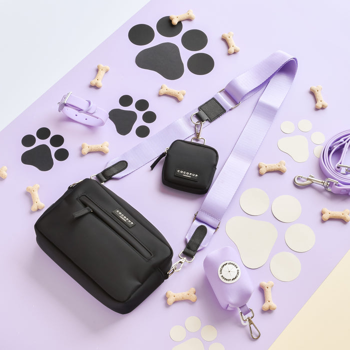 A chic Dog Walking Bag from Cocopup London, featuring a black crossbody design with the NAKD Bag Strap in Parma Violets. The bag showcases purple accessories, including a small pouch, collar, and leash, all set against a lavender background. Additional items displayed are dog bone treats, paw print cutouts, and a waste bag dispenser.