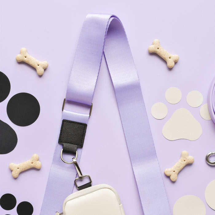 A light purple NAKD Bag Strap - Parma Violets from Cocopup London lies on a pastel purple background, surrounded by bone-shaped dog treats, black and white paw print stickers, and a small cream-colored pouch attached to the strap. The items are aesthetically arranged in a flat lay style.
