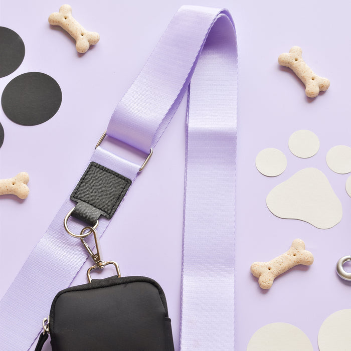 A lilac bag strap with a black pouch from the Cocopup London NAKD Bag Strap - Parma Violets collection is laid out on a light purple surface. Surrounding the strap are dog bone-shaped treats, black and white circles, including a paw print cutout. The arrangement is playful and organized with pet-themed decor.