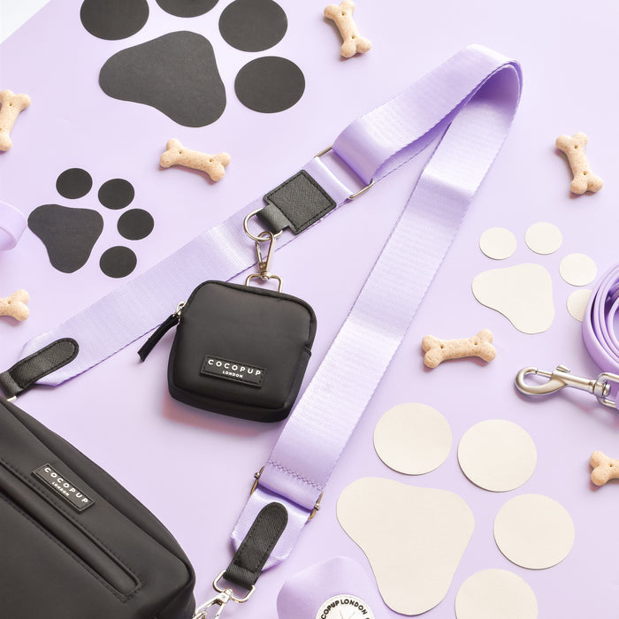 A black crossbody Dog Walking Bag with a matching black pouch, both labeled "Cocopup London," are laid out on a lavender surface. Surrounding the bag are a NAKD Parma Violets leash, bone-shaped dog treats, black paw print cutouts, and beige circular cutouts from the NAKD Parma Violets collection.
