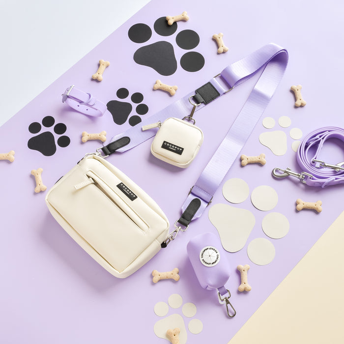 A chic ensemble of lavender dog accessories from Cocopup London's NAKD Parma Violets collection, featuring a leash, collar, waste bag holder, and Dog Walking Bag with the NAKD Bag Strap - Parma Violets, displayed on a light purple surface adorned with black and white paw prints and bone-shaped treats.