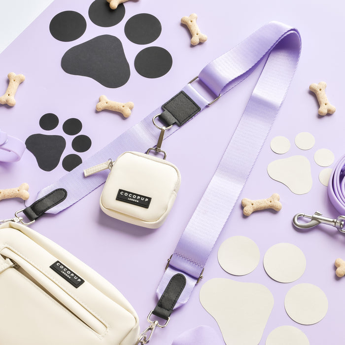 Displayed on a lilac background with black and white paw print cutouts and bone-shaped dog treats scattered around, the dog walking set from Cocopup London includes a beige and purple leash, treat pouch, small bag, and the NAKD Bag Strap from their Parma Violets collection.