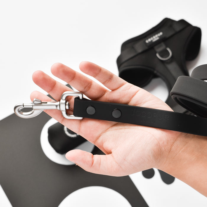 Hand holds black Cocopup London NAKD Lead; black harness and a sleek dog accessory rest in the background.