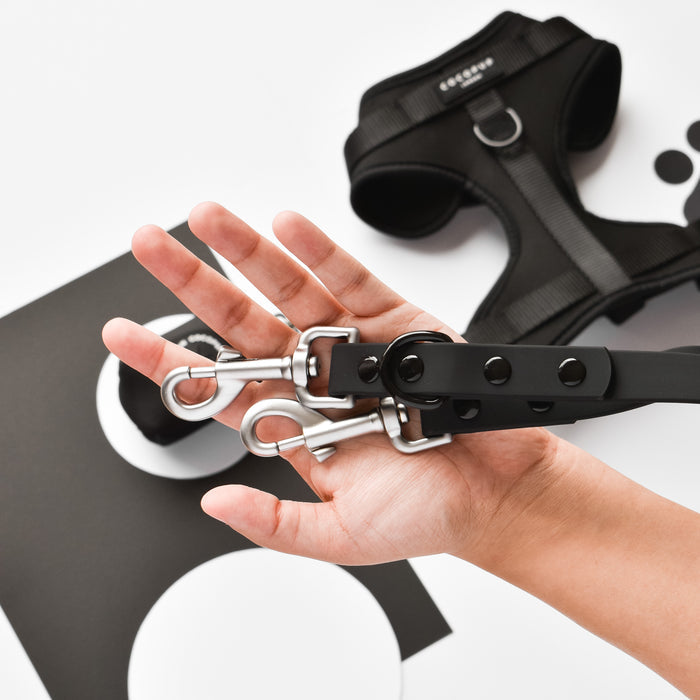 Hand holds metal clasps and black harness; Cocopup London NAKD Lead in liquorice color rests in the background.