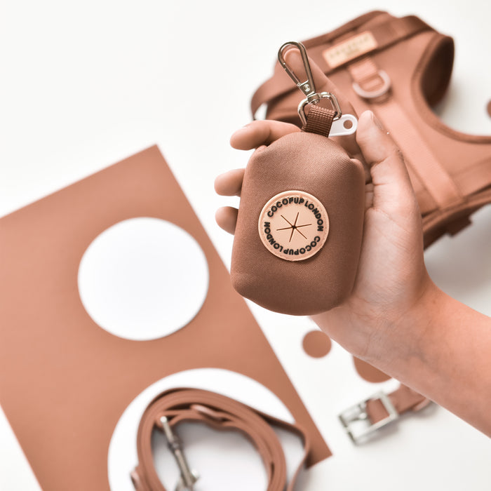 Hand holding brown Cocopup London NAKD poop bag holder, shown with matching brown harness and leash.