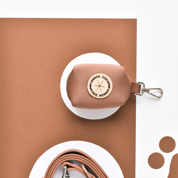 Brown NAKD Poop Bag Holder by Cocopup London with round leather label and metal clip, shown on brown and white surfaces.