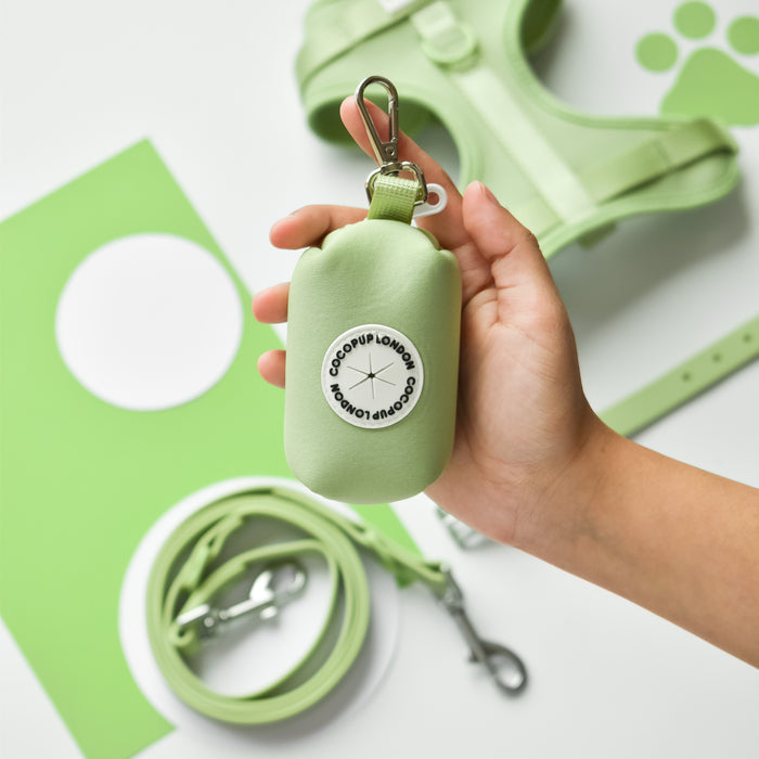 Hand holds a green Cocopup London NAKD Poop Bag Holder next to matching harness and leash set.