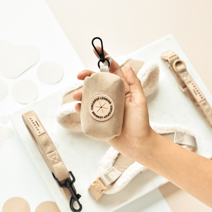 Hand holding a beige Cocopup London Aviator poop bag holder with matching harnesses displayed on a white surface.