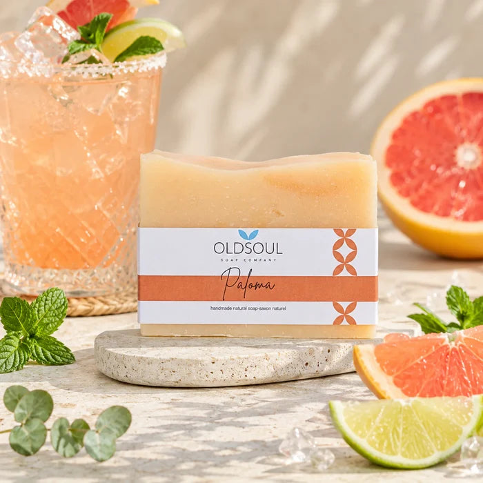 Bar of soap with 'OldSoul Soap Company' packaging on a wooden surface with a cocktail and fruit in the background.