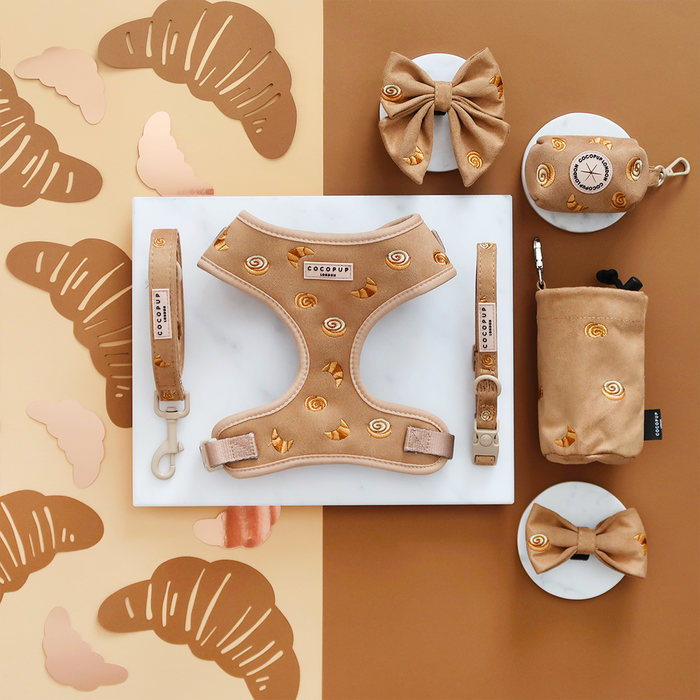 Tan adjustable dog harness with croissant print, shown on a marble and brown croissant-themed background.