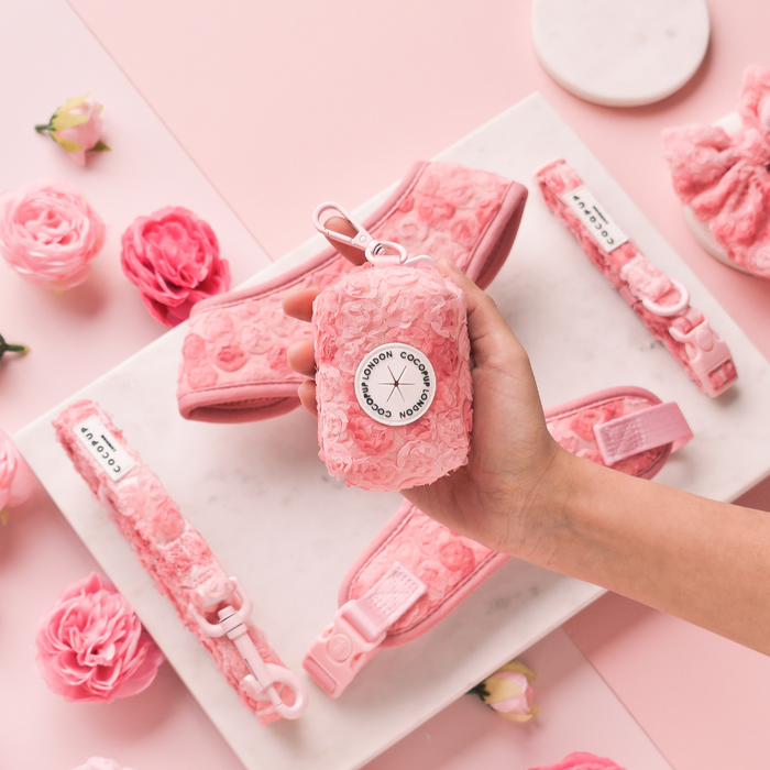 Hand holding a pink Cocopup London Peony Pup Poop Bag Holder with matching accessories and flowers on a pink background.