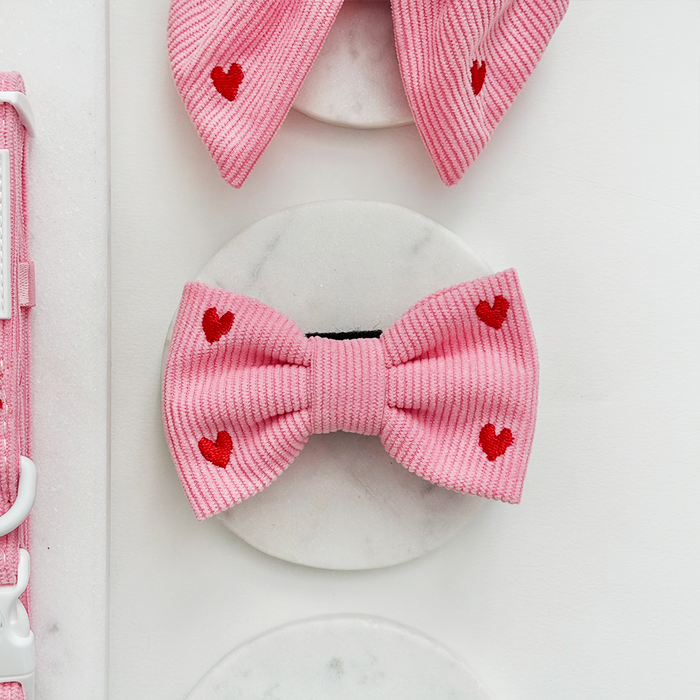 Pink cord bow tie with red heart pattern, displayed on round white marble tiles.