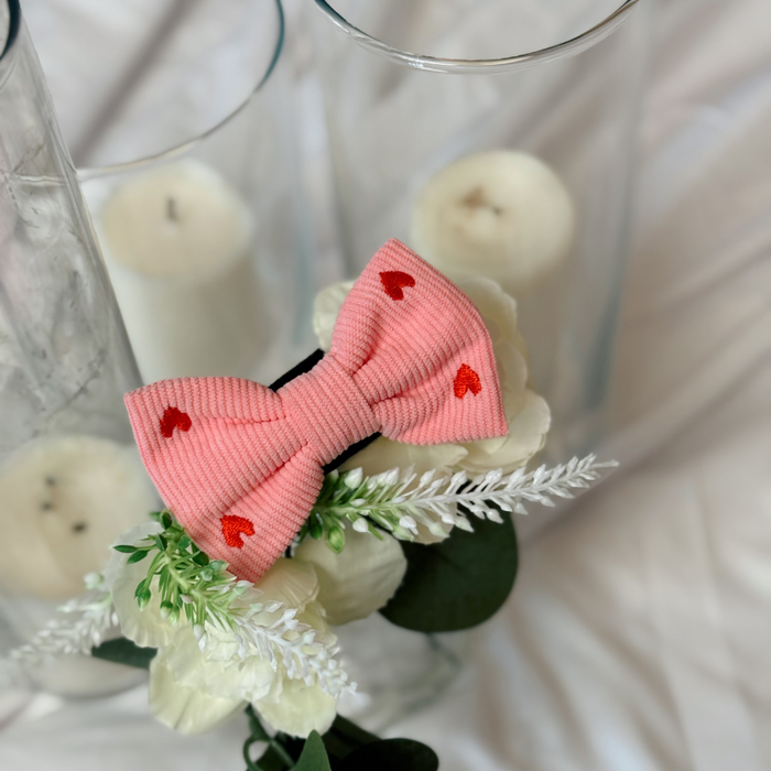 Pink cord bow tie with white heart pattern, displayed alongside white flowers and candles.