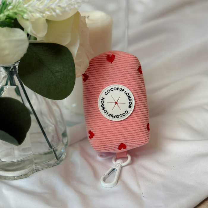 Pink cord poop bag holder with heart pattern on fabric, beside a vase of white flowers.