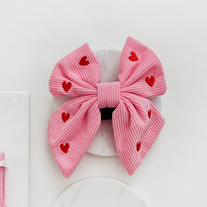 Pink cord sailor bow tie with red hearts pattern from Cocopup London on a white round surface.