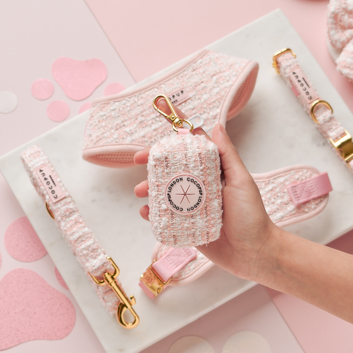 Hand holding a pink textured tweed poop bag holder by Cocopup London, with dog accessories on a marble tray.