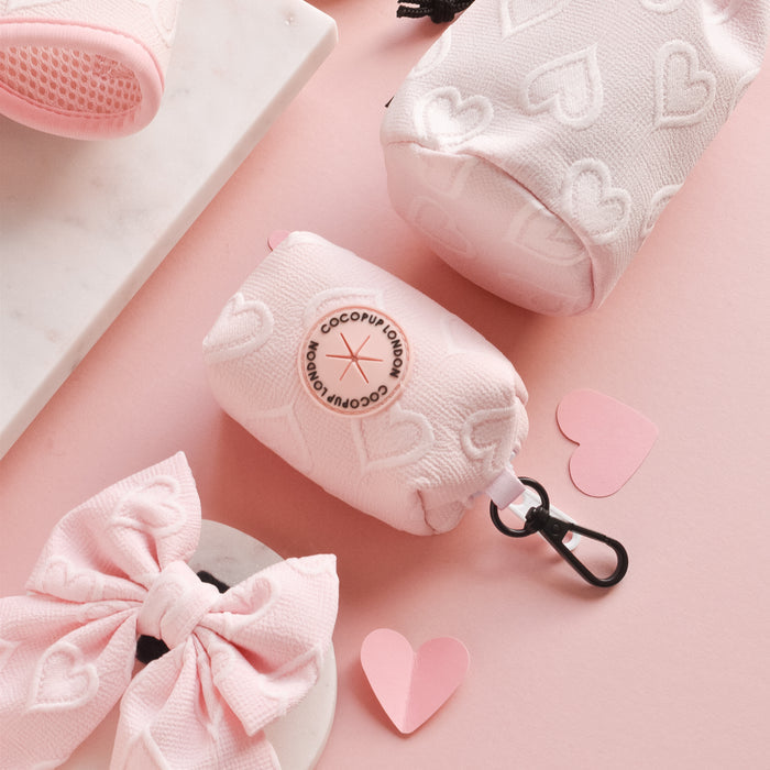Baby pink heart-shaped poop bag holder with a bow, displayed on pink marble with paper hearts.
