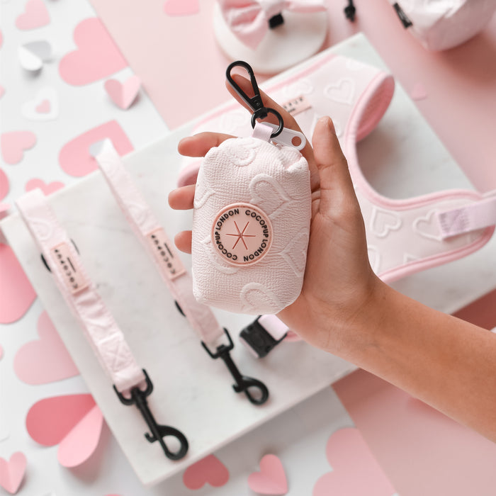 Hand holding pink heart-patterned Luxe Poop Bag Holder by Cocopup London, with matching accessories close by.