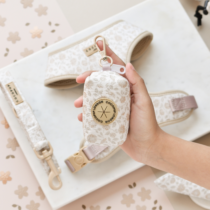 Hand holding a floral-patterned Cocopup London Luxe Poop Bag Holder; matching accessories on a marble tray below.