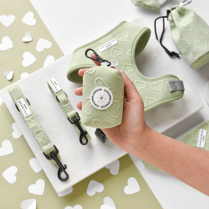 Hand holding a sage green Luxe Poop Bag Holder with heart pattern, alongside matching harnesses and leashes on white surface.