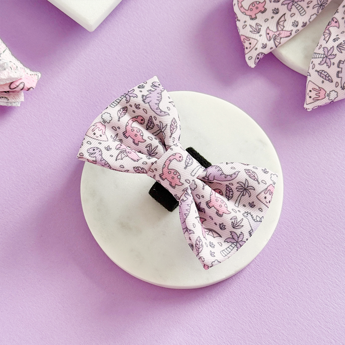 Princess Dino Bow Tie by Cocopup London, pastel dinosaur pattern, displayed on a white platform with lavender background.