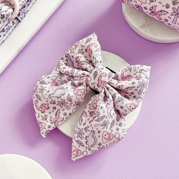 Princess Dino Sailor Bow Tie with pastel dinosaur print by Cocopup London displayed on a dish against a purple background.