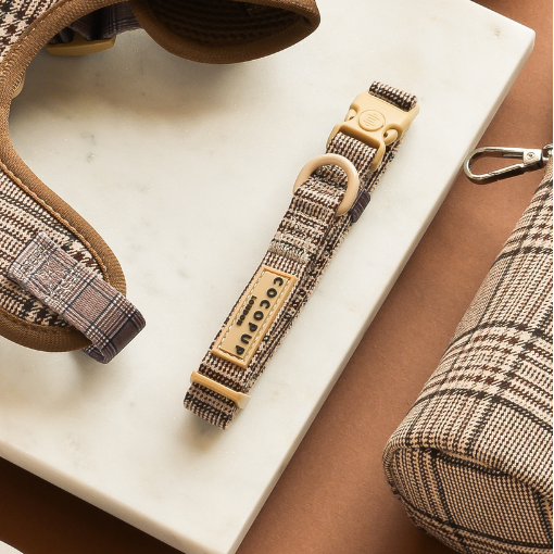 Plaid dog harness, lead, and collar set in brown tones displayed on white marble with a brown background.