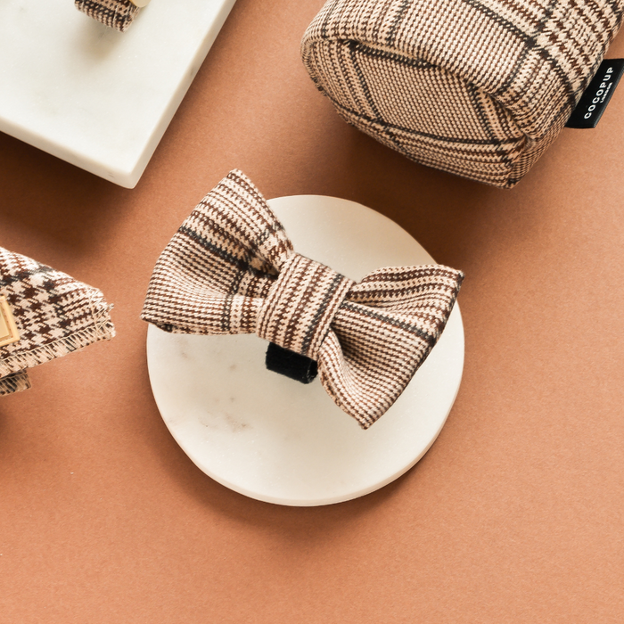 Plaid bow tie and matching tartan accessories displayed on a white dish against a warm brown background.